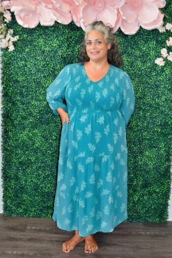 Plus-size woman wearing a teal long-sleeve smocked maxi dress with a subtle leaf print and tiered skirt, standing in front of a floral greenery wall.