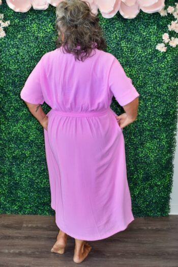 Plus size woman wearing a light lavender button-down shirt dress with a belted waist, smiling and posing in front of a floral and greenery backdrop.