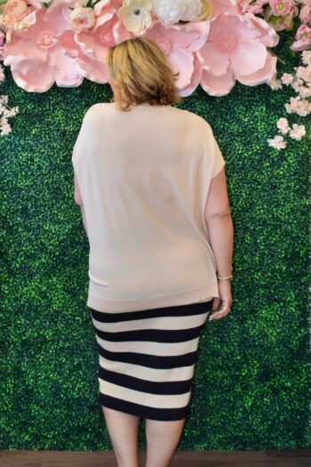 Back view of a plus-size woman wearing a taupe short sleeve knit sweater top, paired with a black and taupe striped skirt, in front of a floral and greenery backdrop.