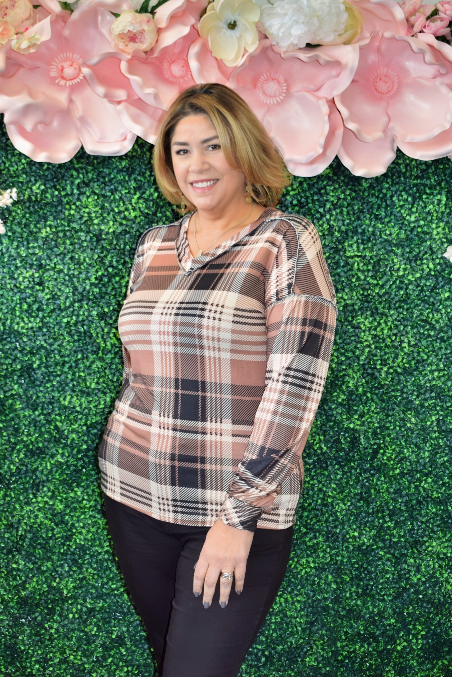 Plus size woman wearing a brown and black plaid long sleeve V-neck top with ruched sleeves, posing in front of a floral green wall at Botticelli Boutique.