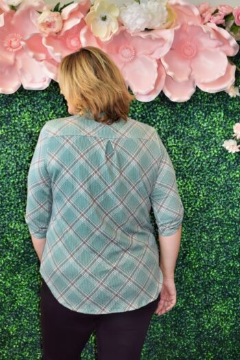 Back view of plus size sage green plaid roll-tab sleeve top with rounded hem and back pleat detail, modeled at Botticelli boutique in Rhode Island.